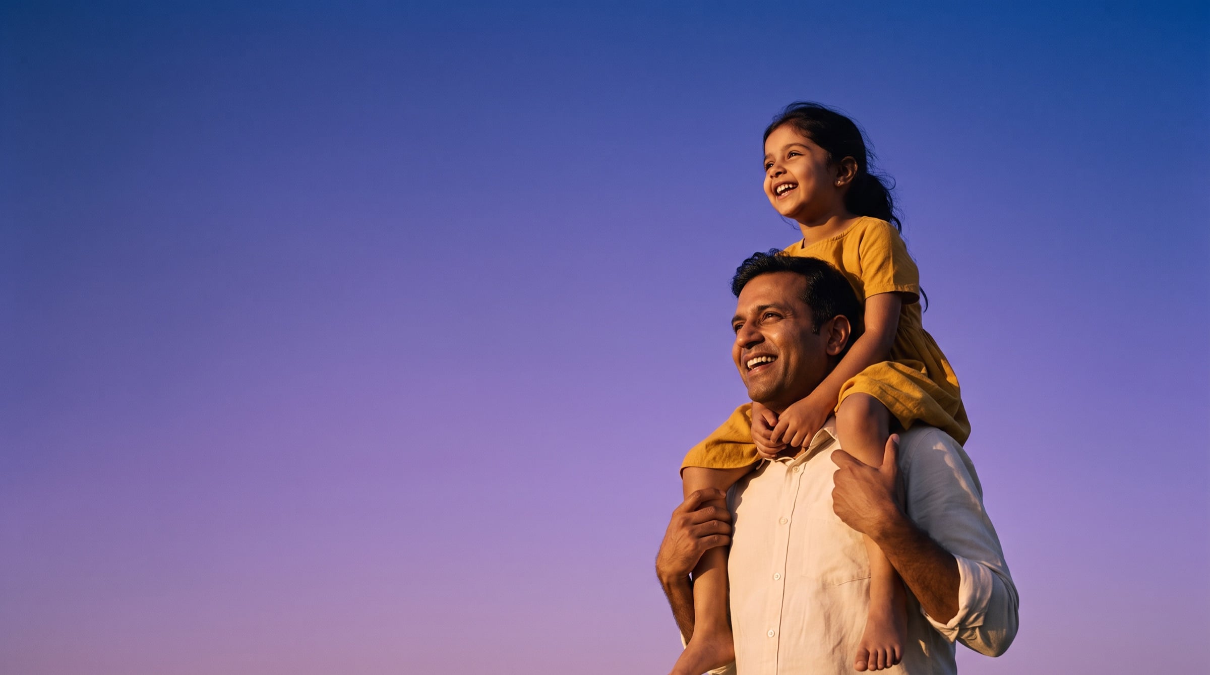 A father carries his laughing daughter on his shoulders against a violet evening sky.