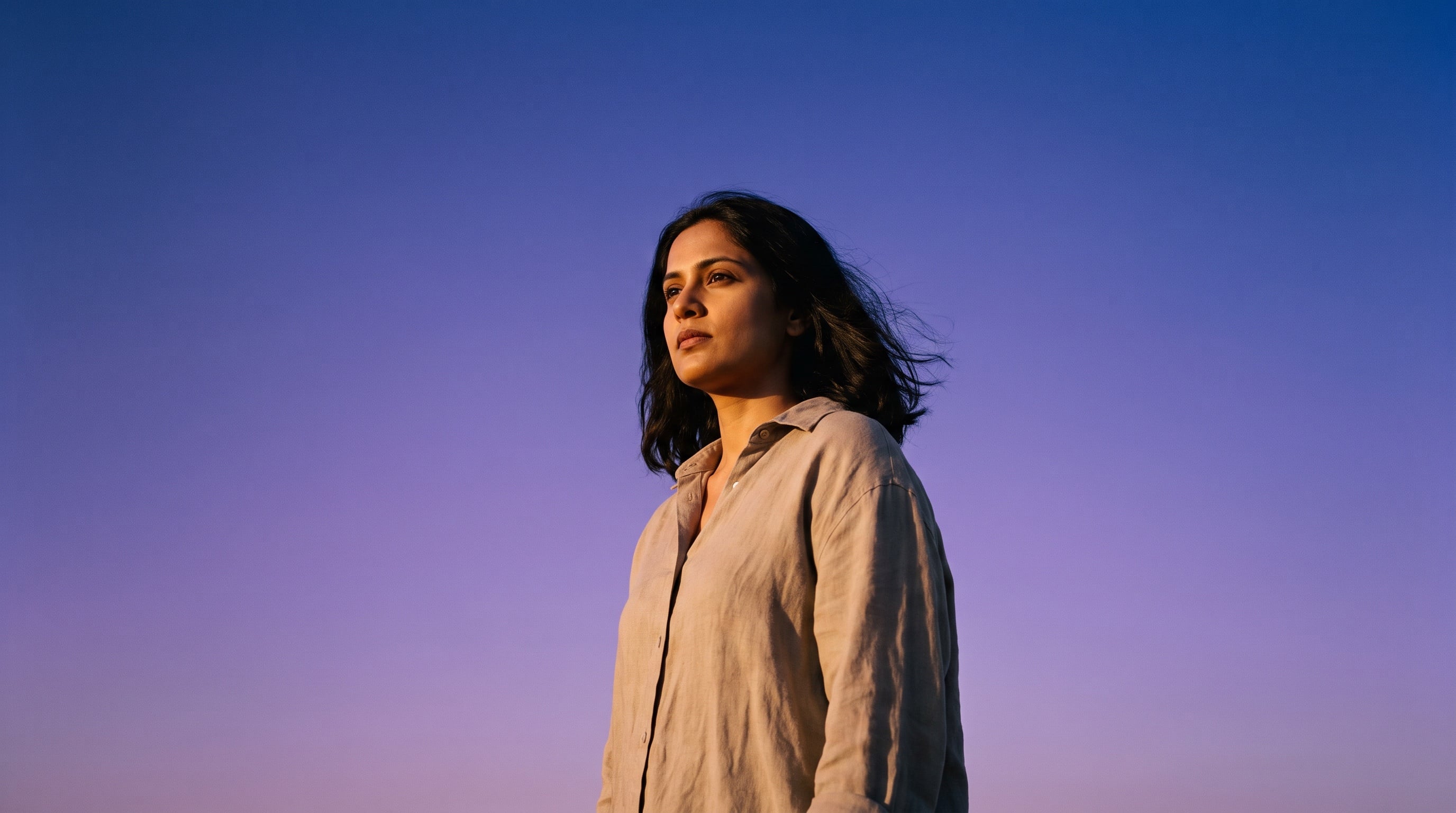 A woman in a linen shirt looks toward the light against a clear violet evening sky.