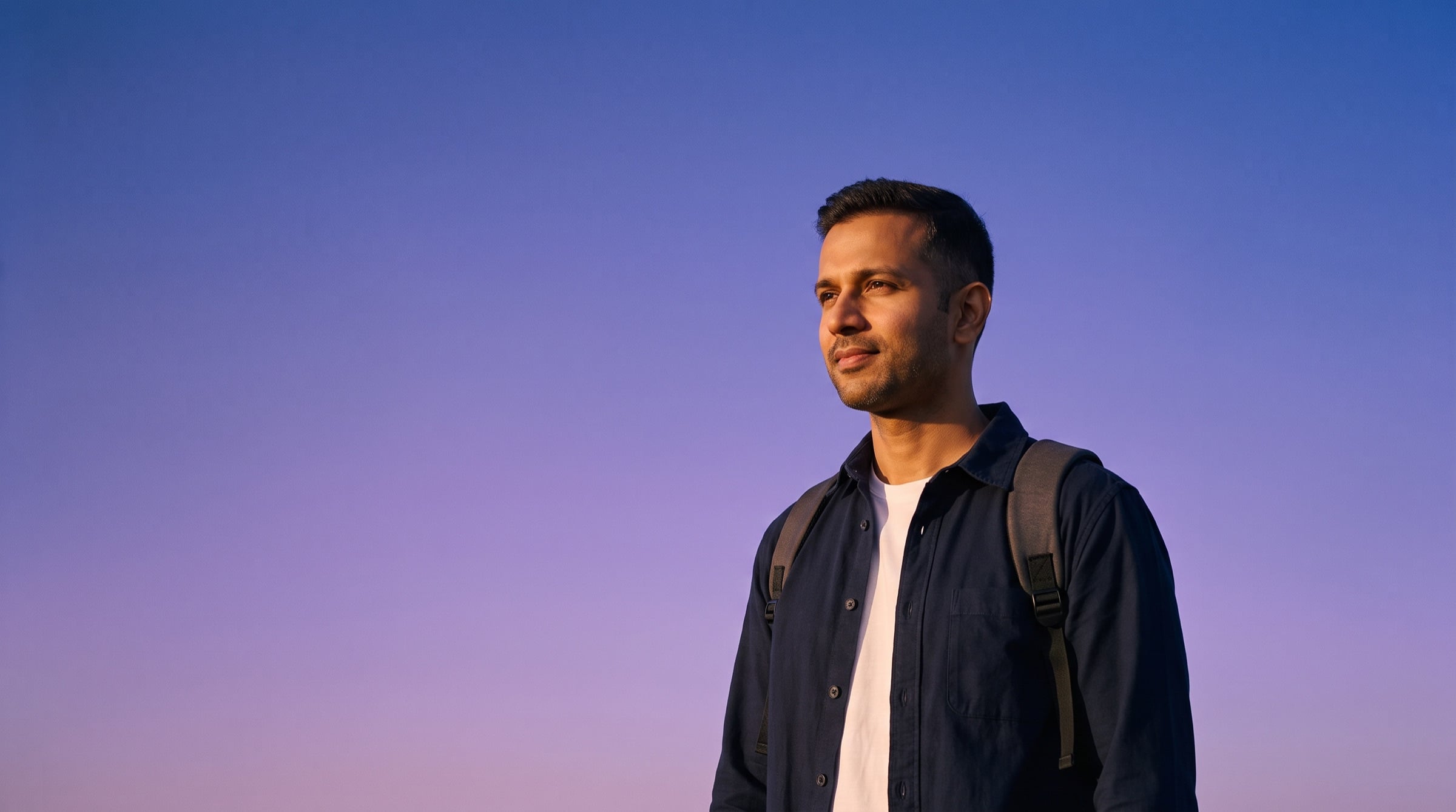 A man in a navy shirt with a backpack looks toward the evening light against a violet sky.